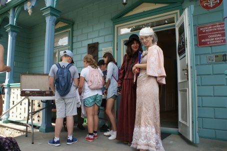 Grupa dzieci i dwie kobiety w starych sukniach na ganku muzeum