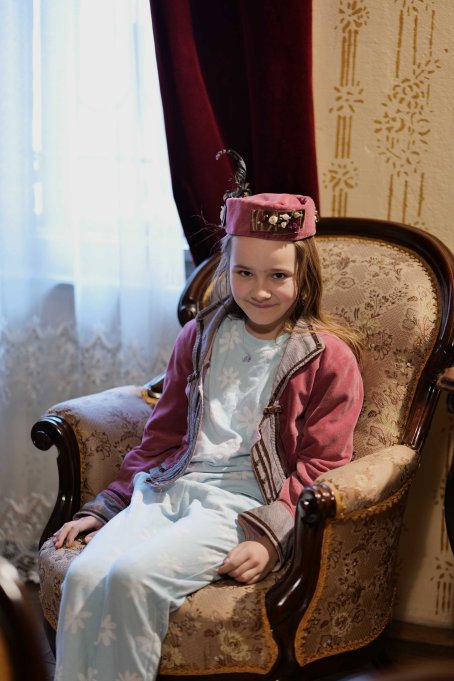 Dziewczynka przebrana w strój z początku XX w. siedzi na fotelu.