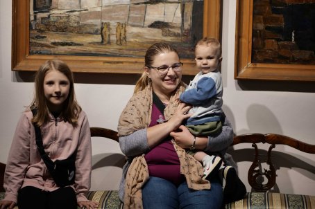 Dziewczynka, obok kobieta z chłopcem, w tle obrazy.