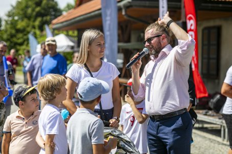 19.	Mężczyzna przemawia przez mikrofon, obok kobieta, dzieci, w tle budynek, ludzie.