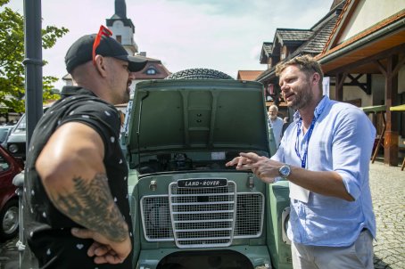 25.	Dwóch mężczyzn rozmawia przed pojazdem, w tle ludzie, budynki.