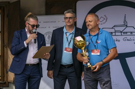 37.	Mężczyzna przemawia przez mikrofon, obok dwóch mężczyzn z plakietkami, jeden z nich trzyma puchar.