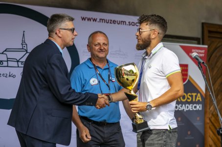 38.	Mężczyzna trzyma w ręce puchar i podaje rękę innemu mężczyźnie, za nimi mężczyzna z plakietką.